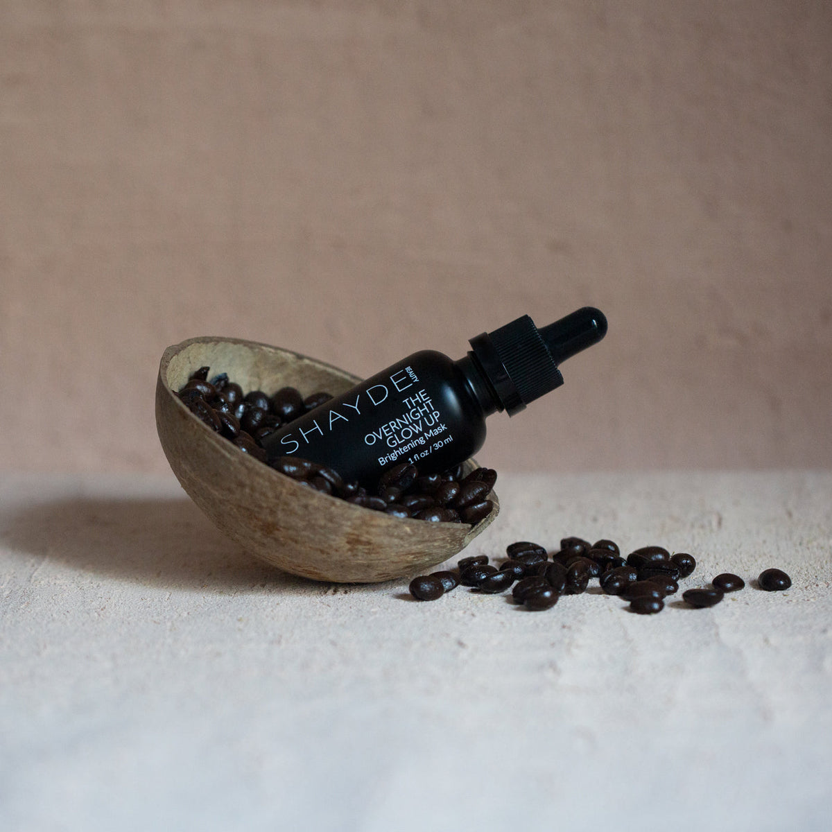 Bottle of OVERNIGHT GLOW UP liquid mask in a coconut shell surrounded by coffee beans.